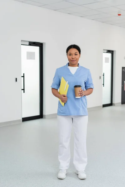 Volle Länge der junge und lächelnde multirassische Krankenschwester mit Papiermappe, Stift und Kaffee zum Mitnehmen Blick in die Kamera im Wartebereich der Blutspendezentrale, Tür auf dem Hintergrund — Stockfoto