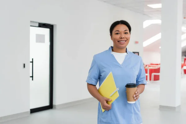 Happy multiracial nurse in blue uniform holding folder with pen, disposable cup with coffee and smiling at camera in waiting area of blood donation center, door on background — Stock Photo