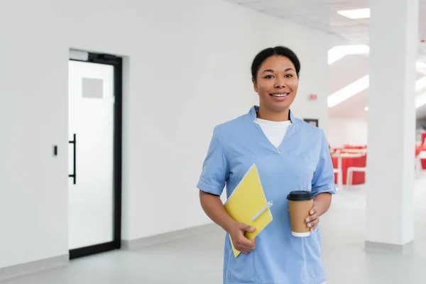 Felice infermiera multirazziale in uniforme blu cartella con penna, tazza usa e getta con caffè e sorridente alla fotocamera nella zona di attesa del centro di donazione di sangue, porta su sfondo — Foto stock