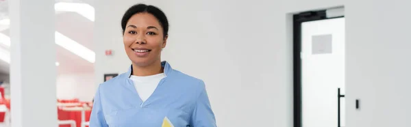 Joyful multiracial healthcare worker in blue uniform looking at camera while standing in waiting area of modern contemporary blood donation center, banner, door on background — Stock Photo