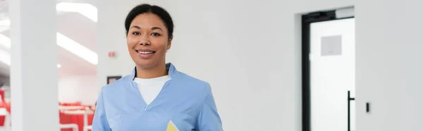 Joyful multiracial healthcare worker in blue uniform looking at camera while standing in waiting area of modern contemporary blood donation center, banner, door on background — Stock Photo