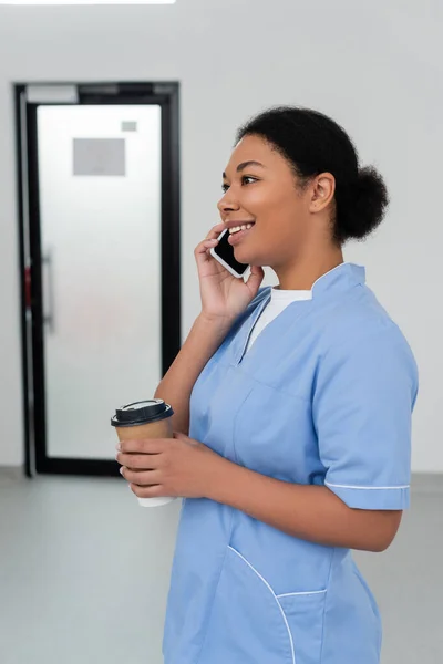 Trabalhador de saúde multirracial alegre em azul uniforme segurando copo de papel com café takeaway e sorrindo durante a conversa no telefone celular no salão do centro de doação de sangue, porta no fundo — Fotografia de Stock