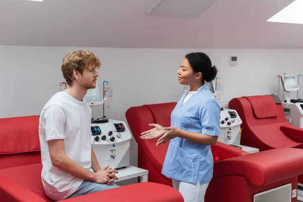 Enfermera multirracial en azul uniforme gesto y hablar con joven donante de sangre pelirroja sentado en silla médica cerca de las máquinas de transfusión en el hospital - foto de stock