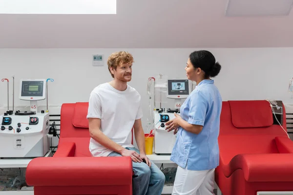 Multiracial nurse in uniform talking to positive redhead volunteer sitting on medical chair near transfusion machines and plastic cup in blood donation center — Stock Photo