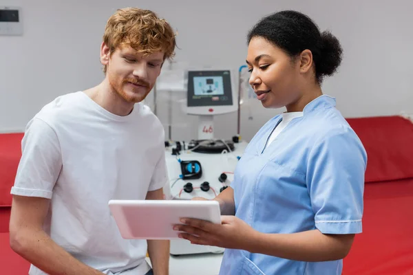 Mitarbeiter des multiethnischen Gesundheitswesens zeigt lächelnden rothaarigen Mann ein digitales Tablet in der Nähe eines verschwommenen Transfusionsgeräts und medizinischer Stühle im Blutspendezentrum — Stockfoto