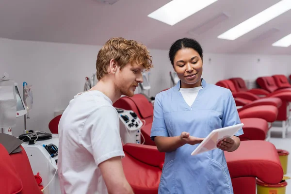 Positive multirassische Krankenschwester in blauer Uniform zeigt auf digitales Tablet in der Nähe junger rothaariger Mann und bequeme medizinische Stühle mit Transfusionsmaschinen in Blutspendezentrum — Stockfoto