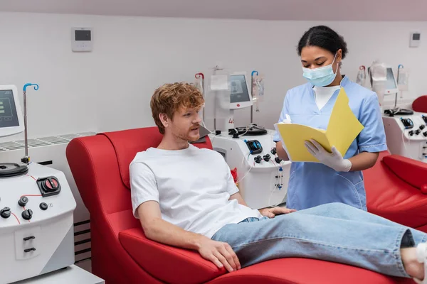 Multiracial nurse in medical mask and blue uniform writing in paper folder near transfusion machine and redhead volunteer on medical chair in blood donation center — Stock Photo