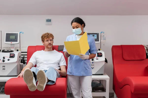 Многорасовый медицинский работник в форме и медицинской маске показывает бумажную папку рыжему волонтеру, сидящему на медицинском стуле возле машины для переливания крови в лаборатории — стоковое фото