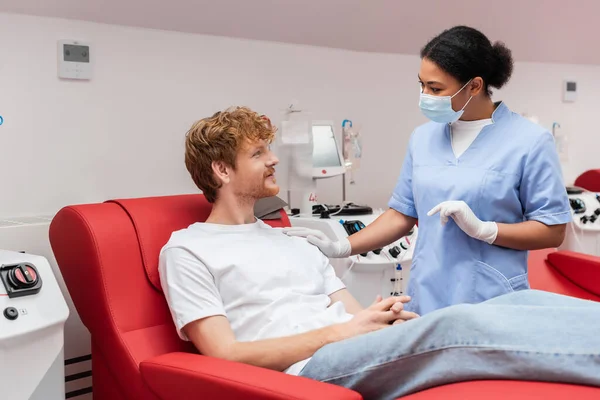 Multiracial nurse in medical mask and latex gloves touching shoulder and talking to redhead blood donor on medical chair near transfusion machine in clinic — Stock Photo