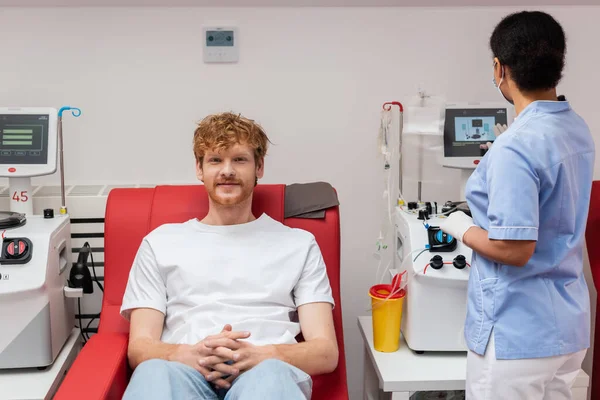 Багаторасовий медсестра в синій уніформі операційної машини переливання поблизу пластикової чашки і рудий доброволець сидить на медичному стільці і дивиться на камеру в центрі донорства крові — стокове фото