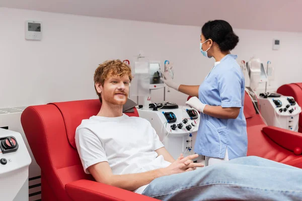 Enfermera multirracial en máscara médica y guantes de látex operando máquina de transfusión cerca del hombre pelirrojo satisfecho sentado en silla cómoda en el centro de donación de sangre - foto de stock