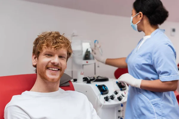Cheerful redhead volunteer looking at camera near multiracial healthcare worker in blue uniform, medical mask and latex gloves operating transfusion machine in blood donation center — Stock Photo