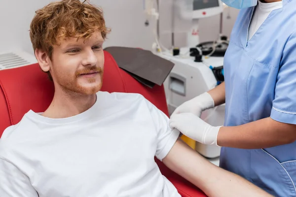 Положительный рыжий донор крови улыбается рядом с многорасовой медсестрой в синей форме и латексных перчатках, сидя на медицинском стуле рядом с размытым аппаратом для переливания крови в центре донорства крови — стоковое фото
