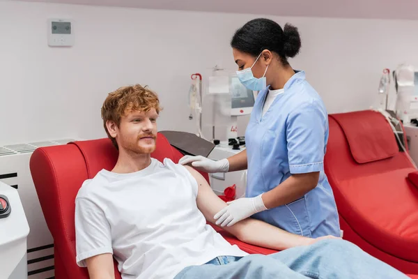 Multiracial nurse in medical mask and latex gloves looking at arm of redhead blood donor sitting on comfortable medical chair near transfusion machine in hospital — Stock Photo
