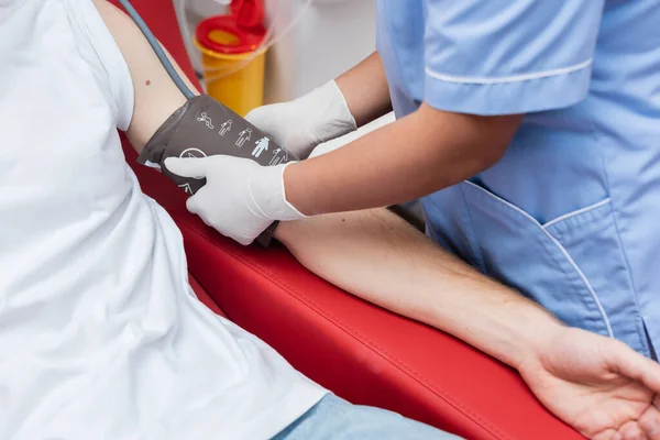 Vista parcial del trabajador sanitario multirracial en uniforme azul y guantes de látex que ponen manguito médico en el brazo del donante de sangre sentado en la silla médica en el hospital - foto de stock