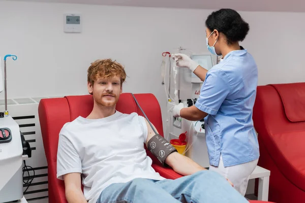 Enfermeira multirracial em máscara médica e luvas de látex montagem transfusão de sangue definido perto de homem ruivo sentado na cadeira médica perto da máquina de transfusão no centro de doação de sangue — Fotografia de Stock