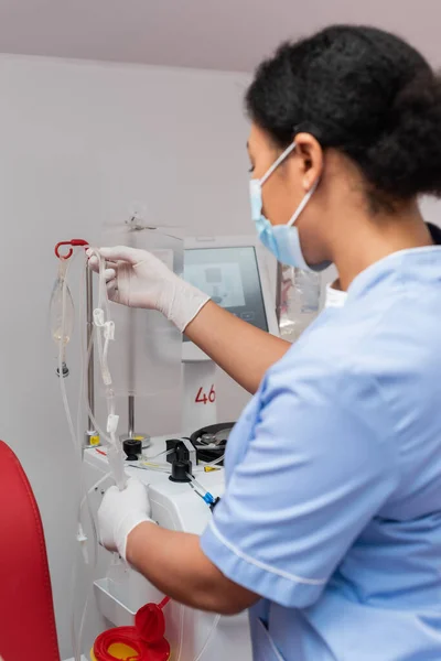 Enfermera multirracial en uniforme azul y guantes de látex que sostienen el equipo de transfusión de sangre cerca del soporte de goteo con bolsas de perfusión en laboratorio, primer plano borroso - foto de stock