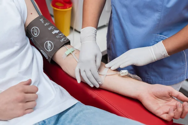Cropped view of multiracial healthcare worker in latex gloves sticking band-aid on arm of volunteer with blood pressure cuff and transfusion set in medical laboratory — Stock Photo