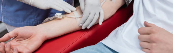Cropped view of multiracial nurse fixing blood transfusion set with band-aid on arm of young volunteer in contemporary blood donation center, banner — Stock Photo
