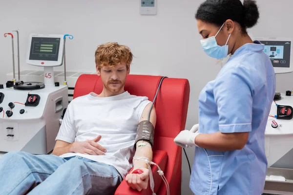 Pelirroja voluntaria con transfusión set apretando pelota de goma mientras está sentada en silla médica cerca de equipo automatizado y enfermera multirracial en máscara médica en centro de donación de sangre - foto de stock