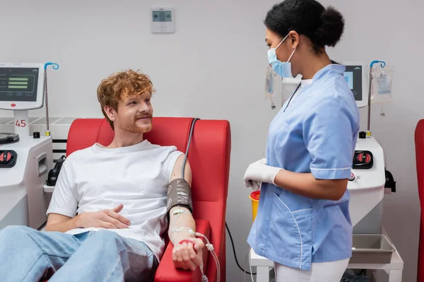 Многорасовая медсестра в медицинской маске и латексных перчатках разговаривает с позитивным рыжим мужчиной с наручниками кровяного давления, сидящим на медицинском стуле возле аппаратов для переливания крови в центре донорства — стоковое фото