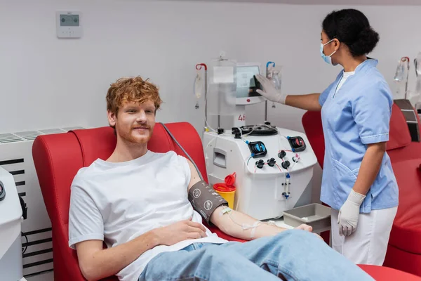 Enfermera multirracial en guantes de látex y máscara médica que opera la máquina de transfusión cerca de voluntario pelirrojo positivo sentado en silla cómoda en el centro de donación de sangre — Stock Photo