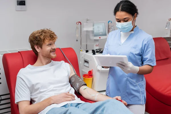Multiracial nurse in medical mask and latex gloves using digital tablet near transfusion machine and redhead donor with blood pressure cuff smiling on medical chair in laboratory — Stock Photo