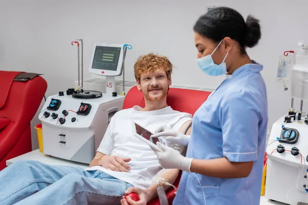 Багаторасова медсестра в синій формі і медична маска стоїть з цифровим планшетом біля усміхненого рудого добровольця на зручному кріслі поруч з автоматизованим обладнанням в центрі донорства крові — стокове фото