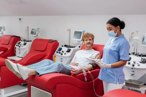 Multirassische Krankenschwester in medizinischer Maske und Latex-Handschuhen mit digitalem Tablet in der Nähe eines rothaarigen Mannes mit Bluttransfusion, der auf einem bequemen ergonomischen Stuhl neben automatisierten Geräten in der Klinik sitzt — Stockfoto