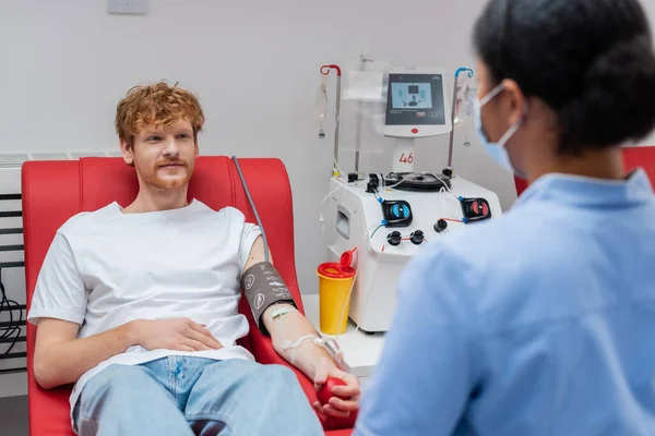 Redhead man in blood pressure cuff holding rubber ball while sitting on medical chair near transfusion machine, plastic cup and multiracial nurse in medical mask on blurred foreground — Stock Photo