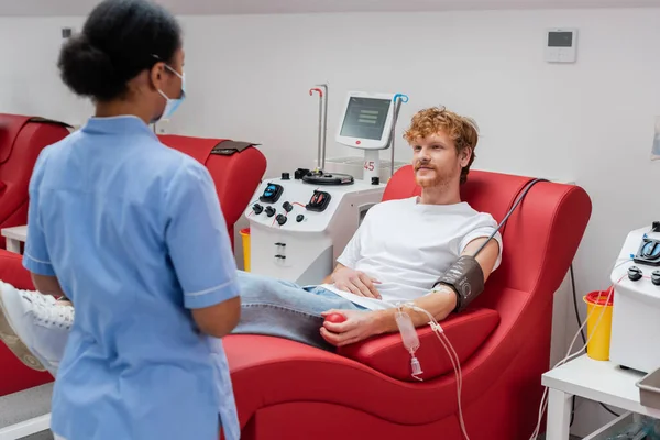 Homem ruivo em transfusão de sangue sentado em cadeira confortável perto de equipamentos automatizados e enfermeira multirracial em uniforme azul e máscara médica no hospital — Fotografia de Stock