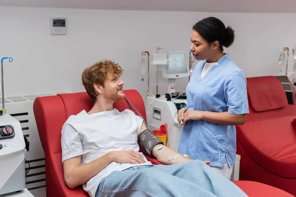 Multiracial nurse in blue uniform talking to redhead donor with blood pressure cuff and transfusion set sitting on medical chair near automated equipment in blood donation center — Stock Photo