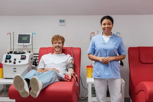 Homem ruivo feliz com manguito de pressão arterial e bola de borracha sentado na cadeira médica perto de enfermeira multirracial, máquina de transfusão e copos de plástico em laboratório, olhando para a câmera — Fotografia de Stock