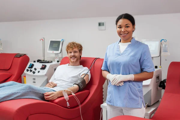 Pelirroja voluntaria con manguito de presión arterial y equipo de transfusión sonriendo a la cámara cerca de la enfermera multirracial en guantes de látex y equipo automatizado en el centro de donación de sangre - foto de stock