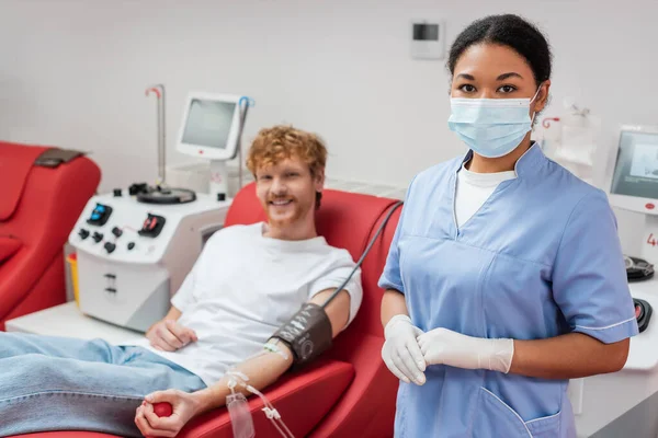 Multiracial healthcare worker in medical mask and latex gloves looking at camera near blurred man with blood transfusion set sitting on ergonomic chair next to transfusion machines in laboratory — Stock Photo