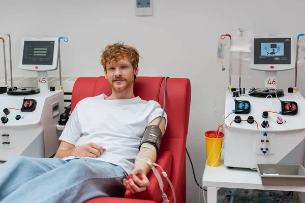 Улыбающийся рыжий мужчина в наборе для переливания крови и манжеты кровяного давления сидя на медицинском стуле и глядя на камеру рядом с автоматизированным оборудованием и пластиковой чашкой в клинике — стоковое фото