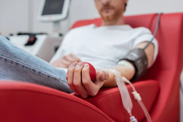 Селективний фокус гумового м'яча в руці обрізаного добровольця з набором переливання, що сидить на зручному медичному кріслі біля машини переливання в центрі донорства крові — стокове фото