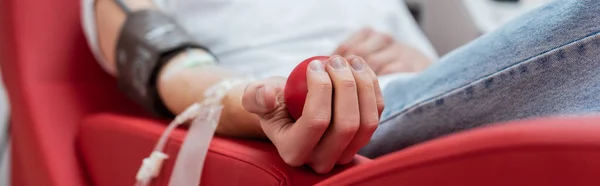 Частичный взгляд молодого волонтера, сидящего на удобном эргономичном стуле и сжимающего резиновый мяч во время медицинской процедуры в центре донорства крови, баннер — стоковое фото