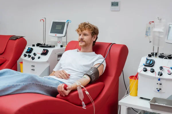 Молодой рыжеволосый донор с наручниками и переливанием крови сидит на эргономичном и удобном медицинском стуле рядом с автоматизированным оборудованием и пластиковыми чашками в клинике — стоковое фото