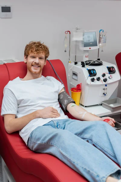 Young and cheerful redhead man with transfusion set sitting on ergonomic medical chair and smiling at camera near modern automated equipment and plastic cup — Stock Photo