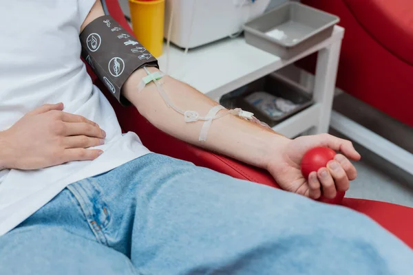 Частичный вид донора с набором для переливания крови, сжимающего резиновый мяч, сидя на удобном стуле рядом с размытым медицинским подносом и пластиковой чашкой в центре донорства крови — стоковое фото