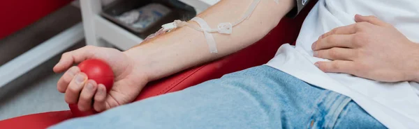 Обрезанный вид волонтера с набором для переливания, сжимающего резиновый мяч, сидя на удобном медицинском стуле в современном центре донорства крови, баннер — стоковое фото