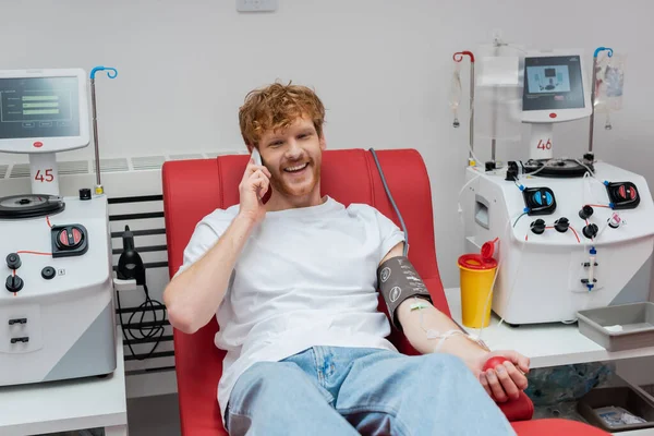 Freudig rothaarige Freiwillige im Transfusionsset auf dem Behandlungsstuhl sitzend und während des Blutspendeprozesses in der Nähe moderner automatisierter Geräte und Plastikbecher im Labor auf dem Smartphone sprechend — Stockfoto