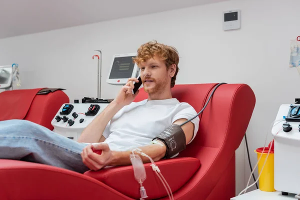 Rotschopf-Spender mit Transfusionsset sitzt auf Medizinerstuhl und spricht auf Smartphone in der Nähe moderner automatisierter Geräte und Plastikbecher im Blutspendezentrum — Stockfoto