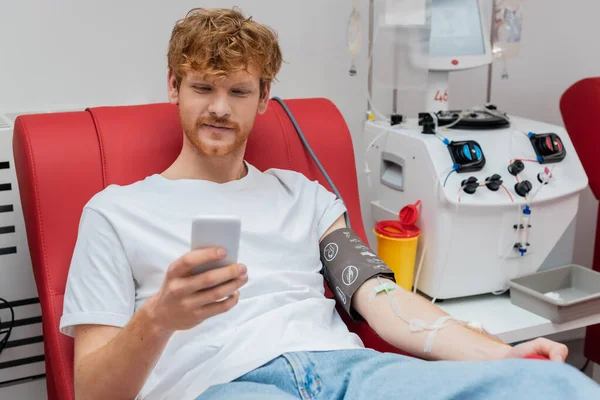 Рудий доброволець з набором переливання, що переглядає Інтернет на смартфоні, сидячи на медичному стільці біля пластикової чашки та машини переливання під час пожертвування крові в клініці — стокове фото