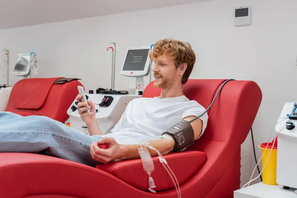 Ruiva e homem sorridente navegando na internet no telefone celular em cadeira médica perto de máquinas de transfusão automatizadas e copo de plástico no centro de doação de sangue — Fotografia de Stock