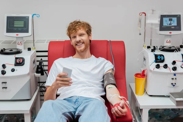 Весёлый рыжий мужчина с наручниками и набором для переливания крови, держащий мобильный телефон, сидя на медицинском стуле рядом с автоматизированным оборудованием и пластиковой чашкой в лаборатории — стоковое фото