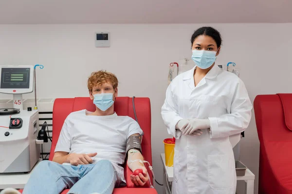 Médecin multiracial dans un masque médical près d'un volontaire roux avec brassard de pression artérielle et ensemble de transfusion assis sur une chaise médicale à côté de l'équipement automatisé et tasse en plastique en laboratoire — Photo de stock