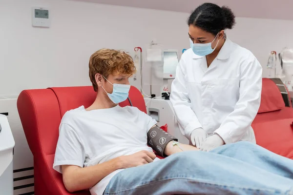 Médico multirracial en bata blanca y máscara médica mirando pelirroja voluntario con manguito de presión arterial sentado cerca de equipos automatizados en el centro de donación de sangre - foto de stock