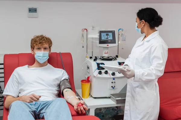 Рыжеволосый донор крови в медицинской маске и манжете кровяного давления, смотрящий на камеру на медицинском стуле рядом с многорасовым врачом с цифровым планшетом, аппаратом для переливания крови и пластиковой чашкой в лаборатории — стоковое фото